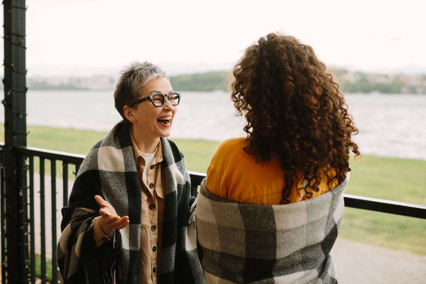 Two women wrapped in blankets laughing together on a porch by the water