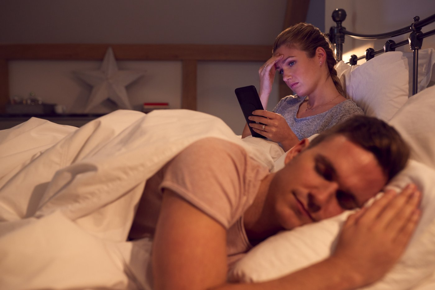 A woman awake in bed looking at her phone while her partner sleeps